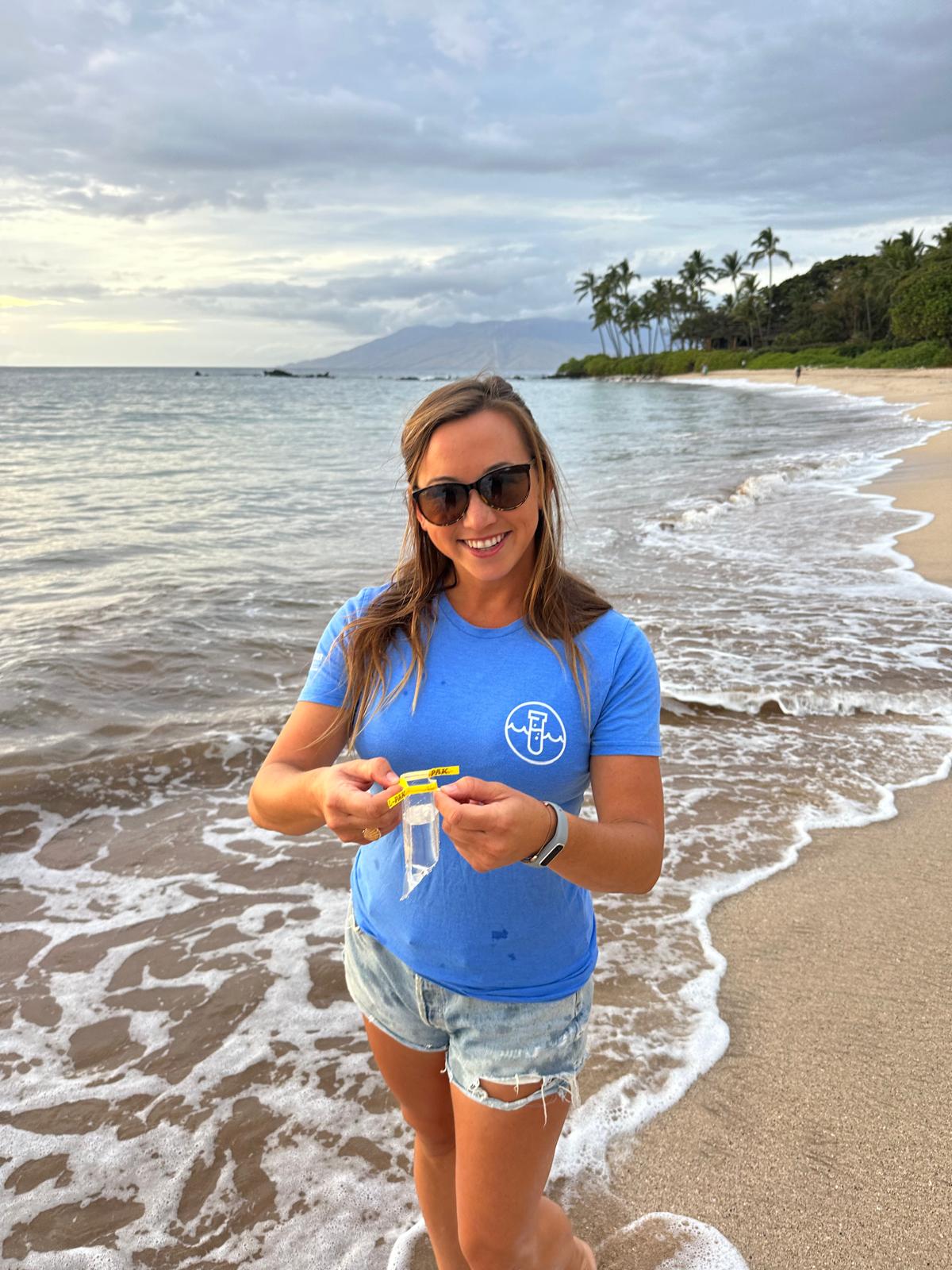 A woman in a blue shirt holding up a water sample on a beach. 