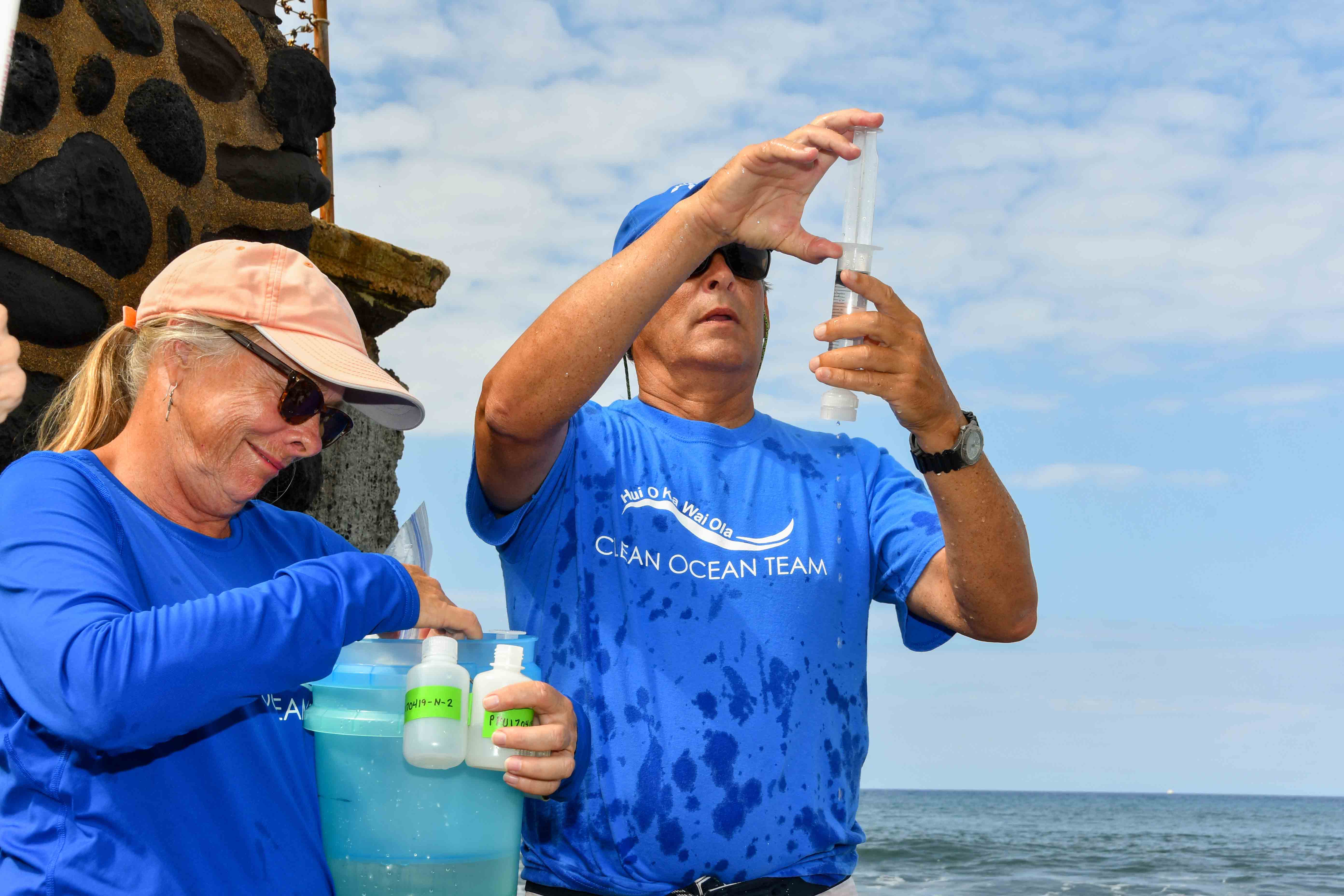 A man wearing a blue hat and blue shirt holds up and examines a tube containing a water sample.